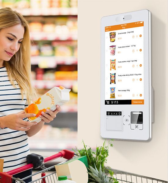 A shopper holding a juice carton next to a wall-mounted self-checkout kiosk in a supermarket